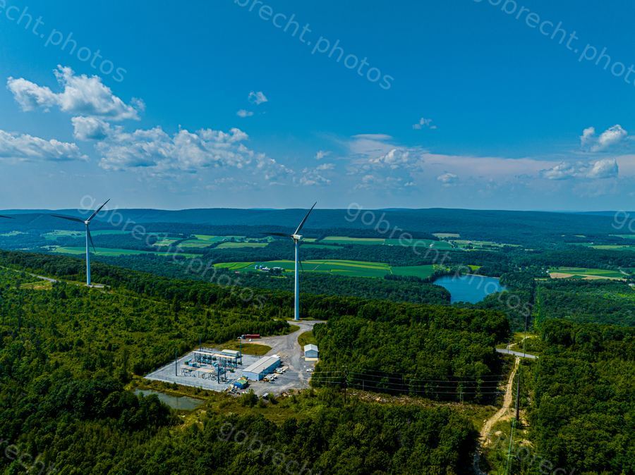 Two wind turbines on a forested hilltop with agricultural valleys and a lake visible in afternoon sunlight.