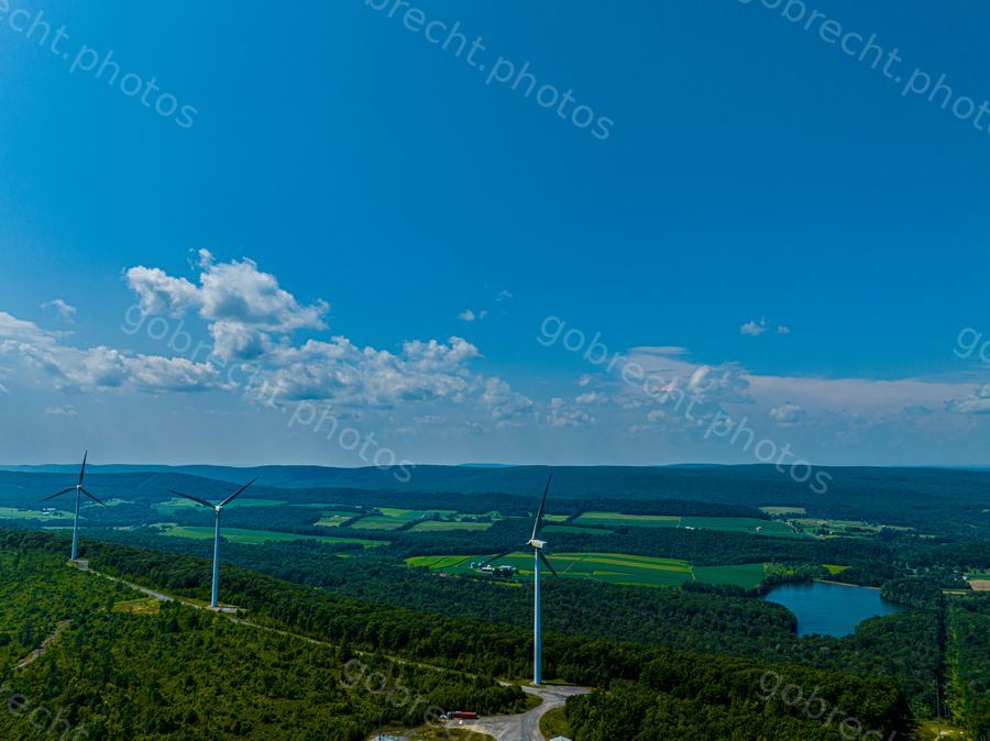 Wind turbines on forested hillside overlooking agricultural valley with lake under blue sky and white clouds.