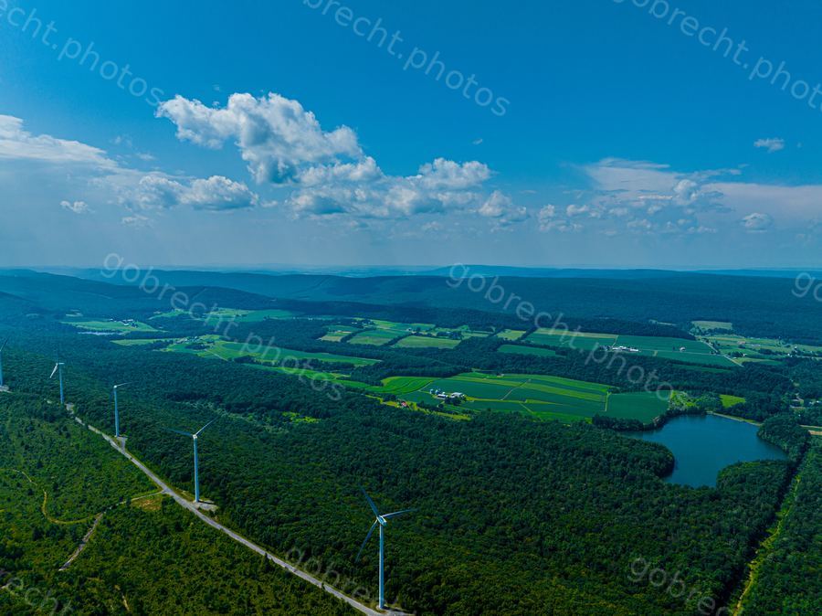 Aerial view of forested landscape with wind turbines, agricultural fields, and a lake under afternoon sunlight.