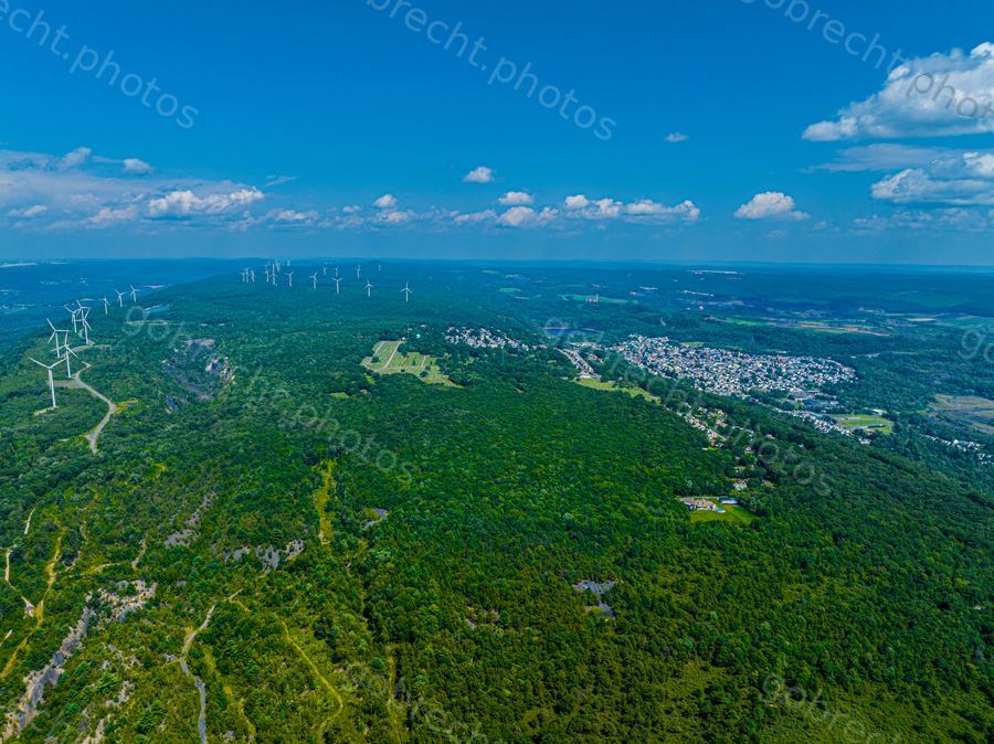 Aerial view of forested landscape with wind turbines, residential areas, and winding roads under afternoon sunlight.