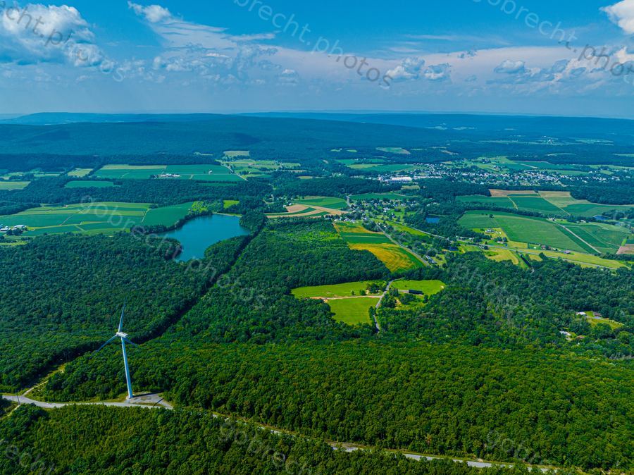 Aerial view of forested landscape with wind turbine, lake, and agricultural fields under afternoon light.