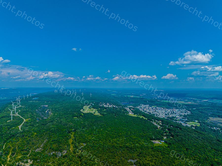 Aerial view of forested landscape with wind turbines, town, and blue sky with white clouds in afternoon light.