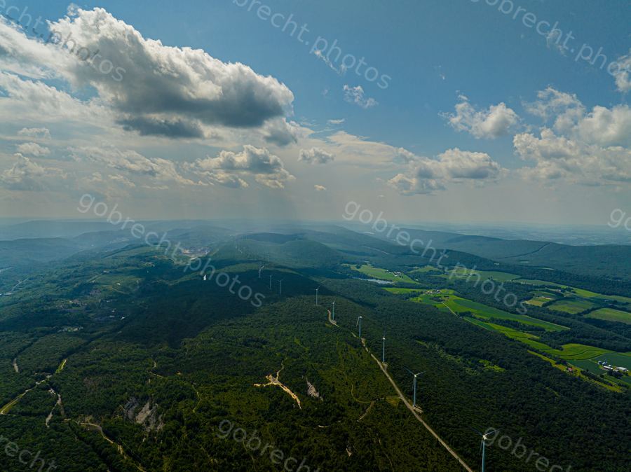 Aerial view of forested rolling hills with transmission towers and cleared agricultural areas under afternoon sky.