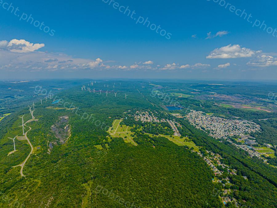 Aerial view of forested hillside with wind turbines, residential areas, and agricultural fields under afternoon sunlight.