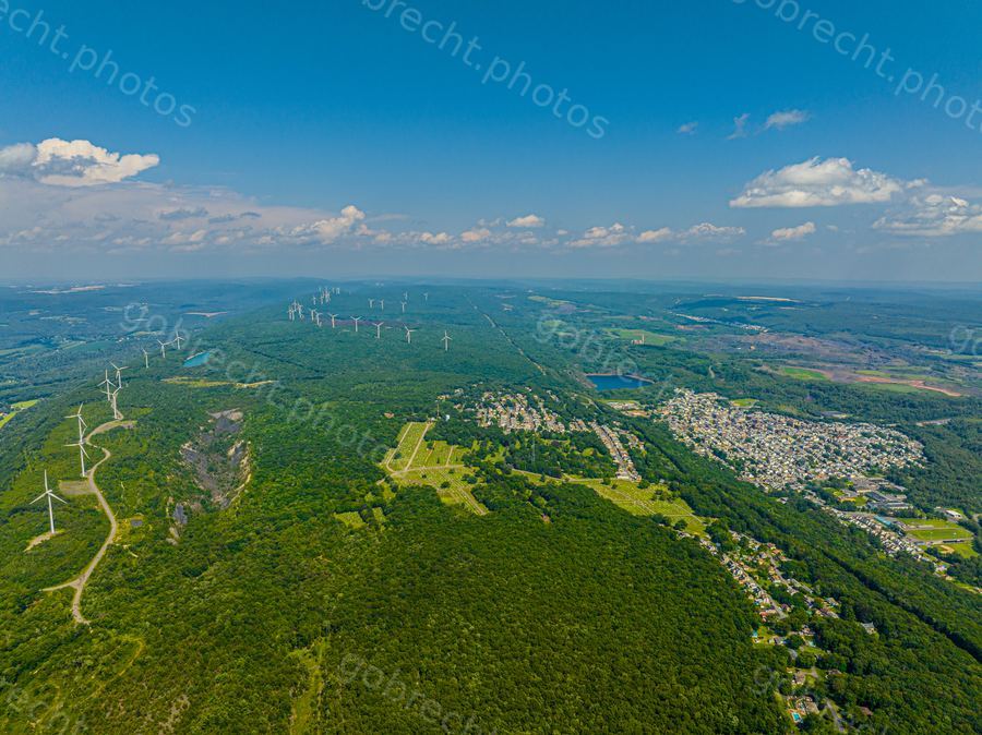 Aerial view of forested mountainous terrain with wind turbines, residential areas, and afternoon light under blue sky.